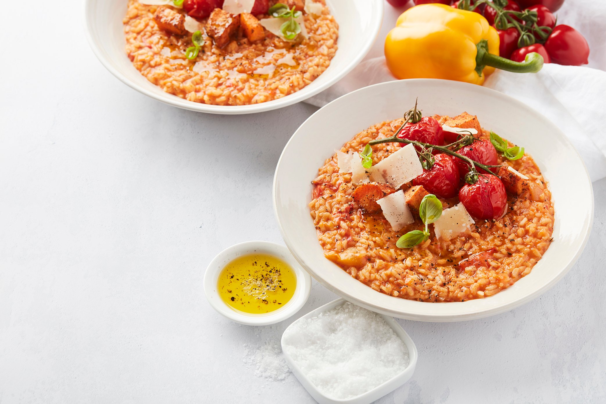 Een culinaire foodfoto van een kom risotto met paprika en zoete aardappel, gegarneerd met geroosterde Frieda tapastomaten, schilfers Parmezaanse kaas en verse basilicum.