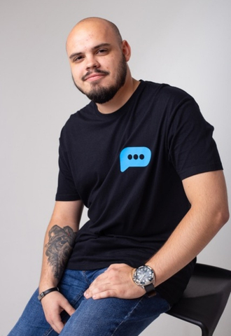 Mann mit schwarzem T-Shirt mit blauem Chat-Symbol sitzt auf Hocker und lächelt leicht.