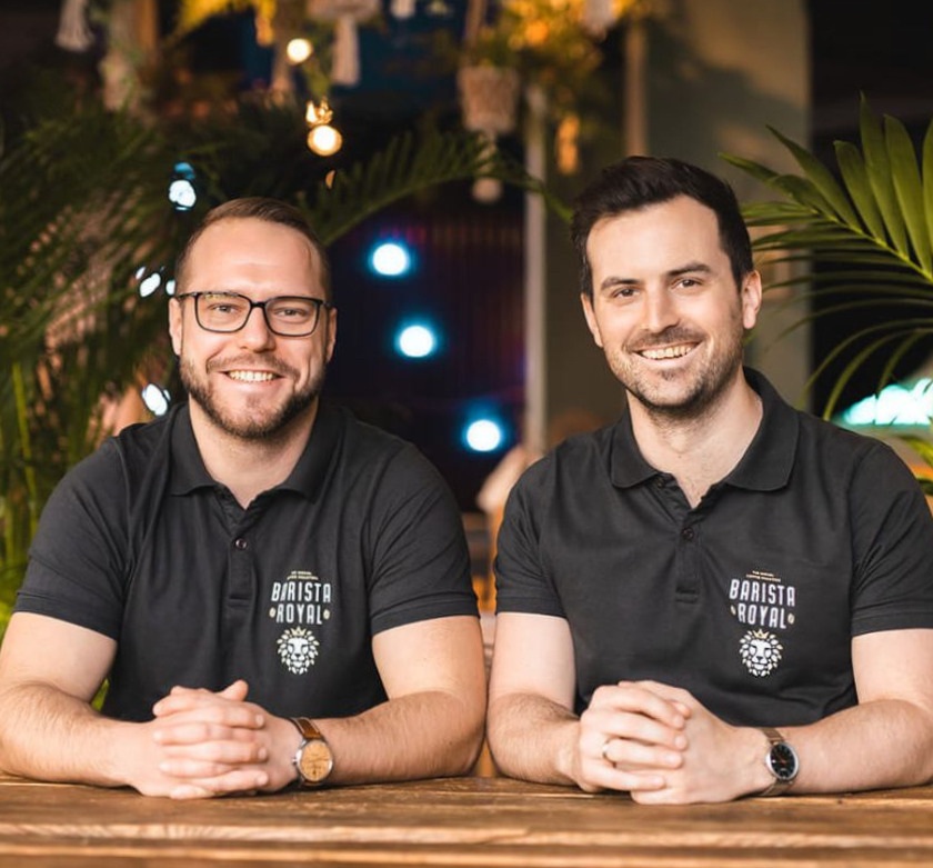 Zwei Männer sitzen nebeneinander, tragen Barista Royal Polohemden und lächeln an einem Holztisch.