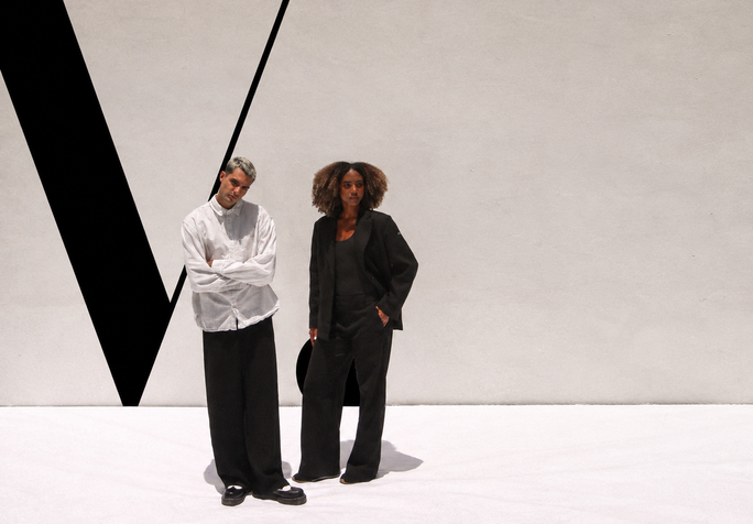 Two people standing in front of a large painted VOUS V