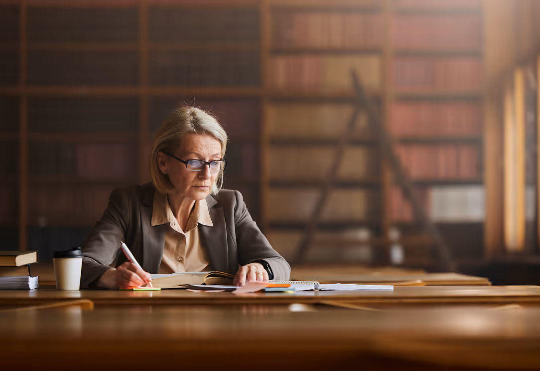Middle-aged woman with glasses writing notes in a book at a large wooden table in a quiet library.