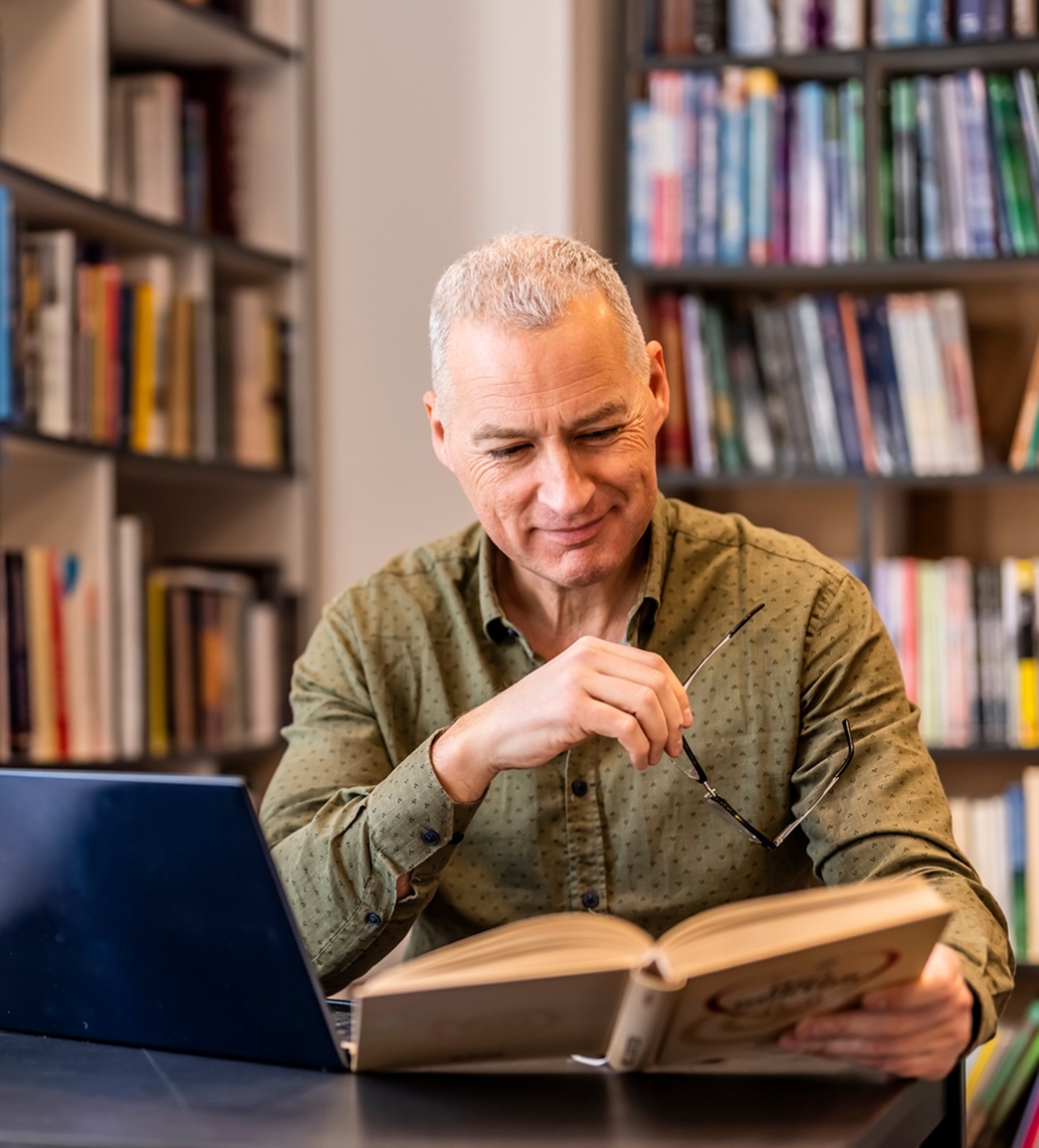 Middle-aged woman with glasses writing notes in a book at a large wooden table in a quiet library.