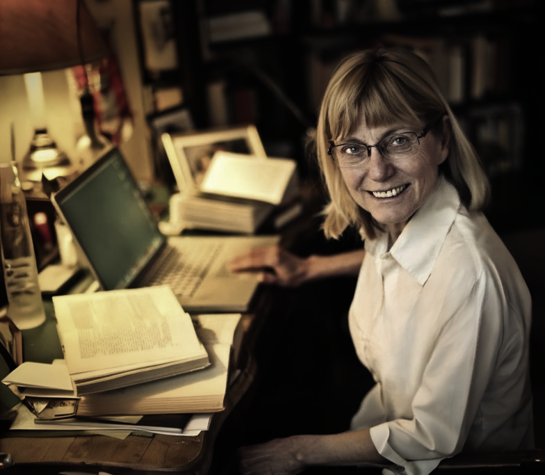 Middle-aged woman with glasses writing notes in a book at a large wooden table in a quiet library.