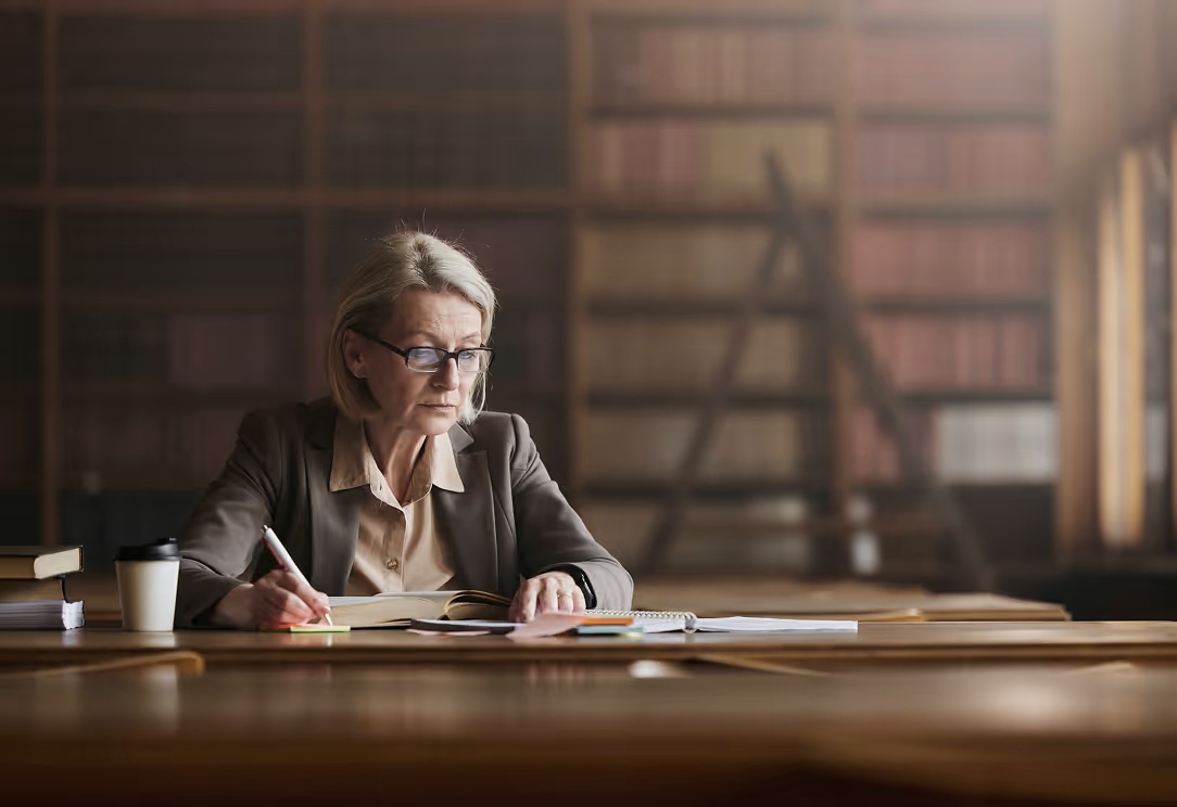 Middle-aged woman with glasses writing notes in a book at a large wooden table in a quiet library.