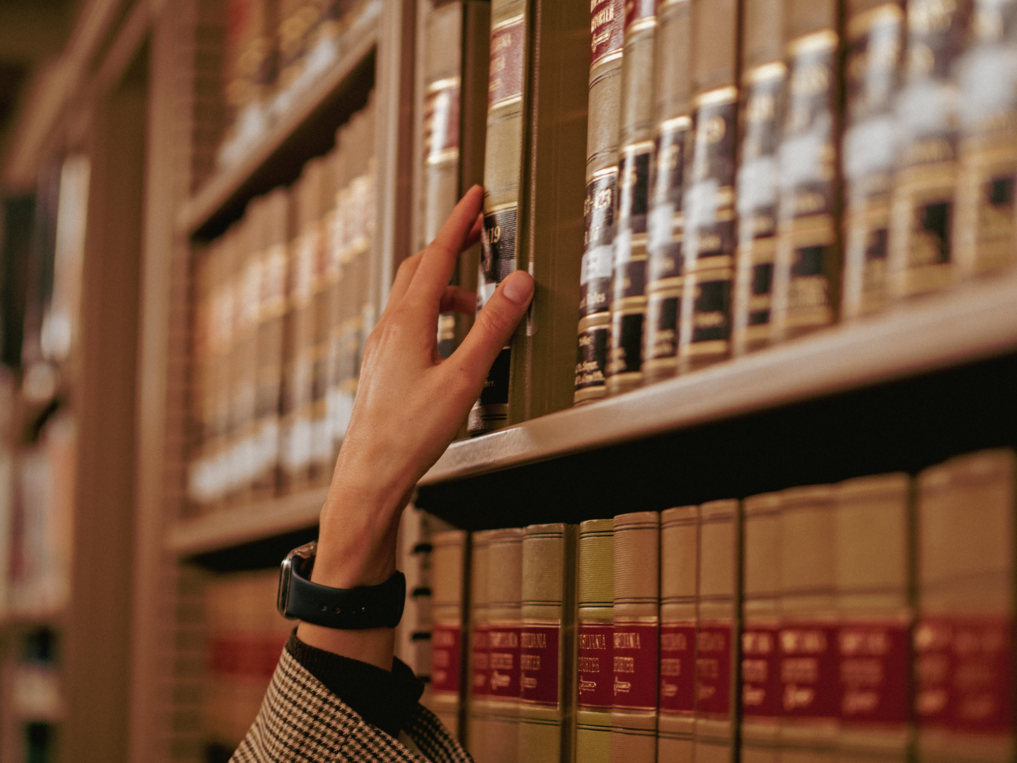 Middle-aged woman with glasses writing notes in a book at a large wooden table in a quiet library.