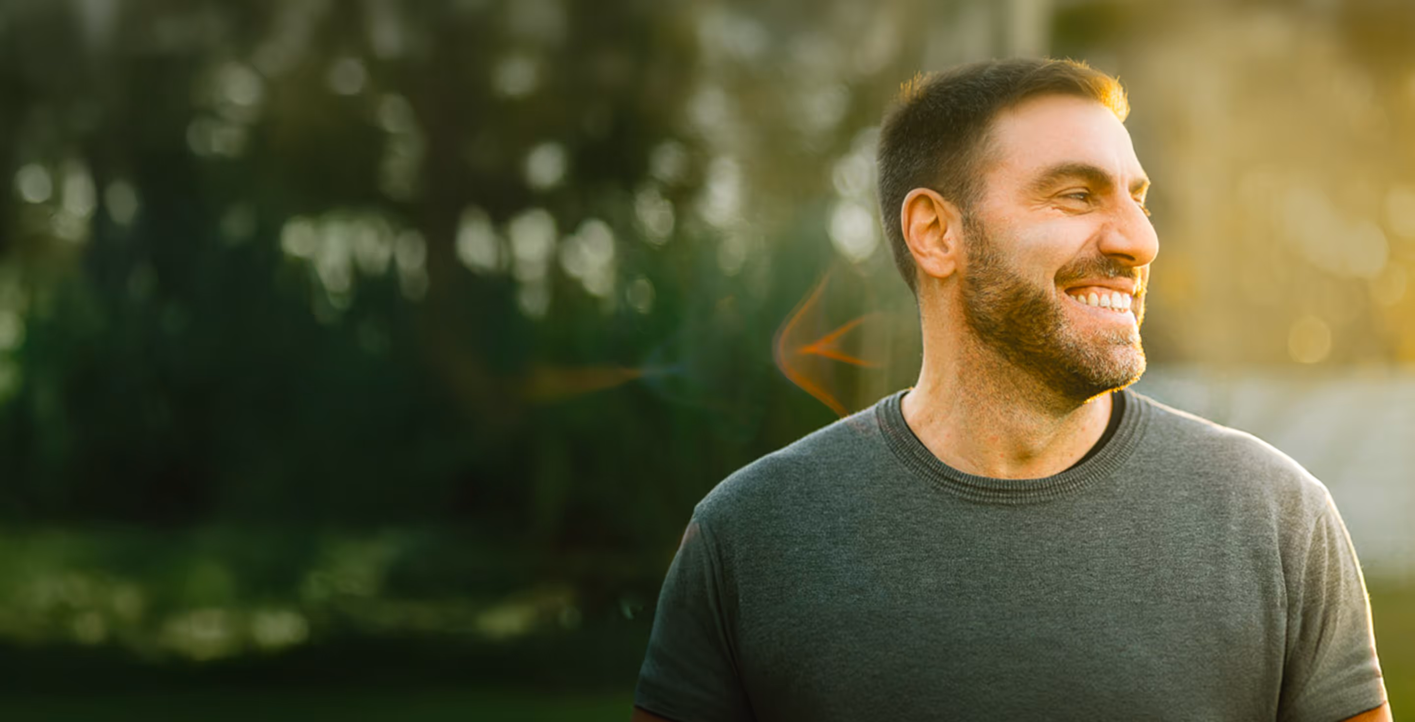 Smiling bearded man in gray shirt looking right with blurred greenery and soft sunlight background.