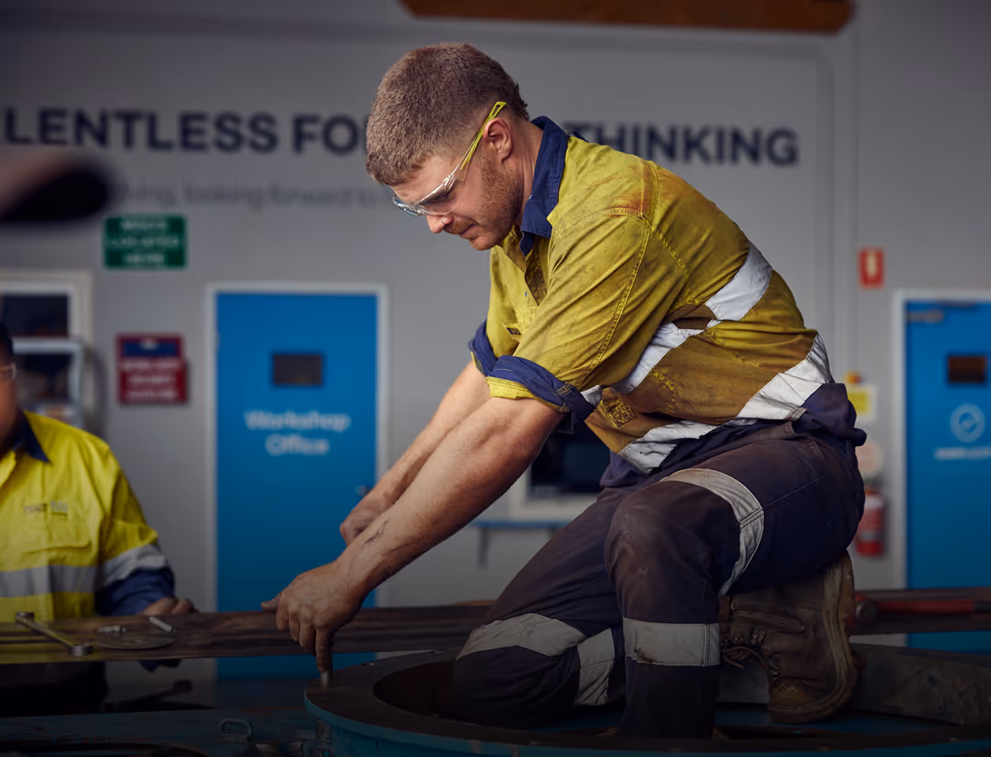Worker in high-visibility clothing and safety glasses kneeling and using a tool on machinery in an industrial workshop.