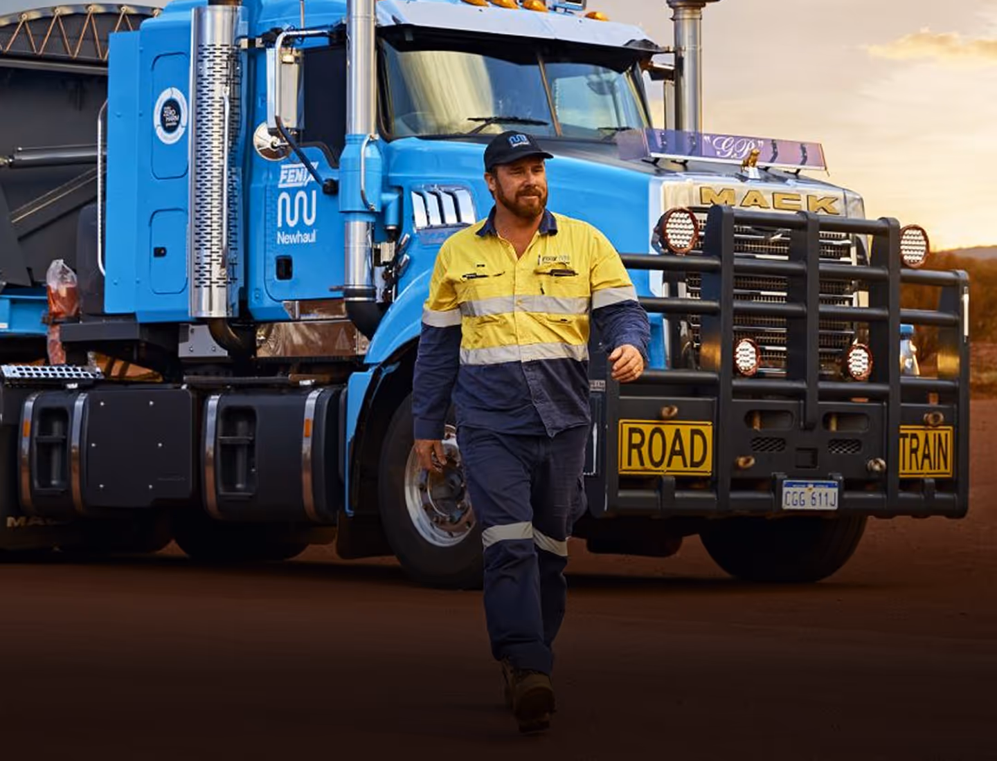 Man in high-visibility workwear walking in front of a blue Mack road train truck at sunset.