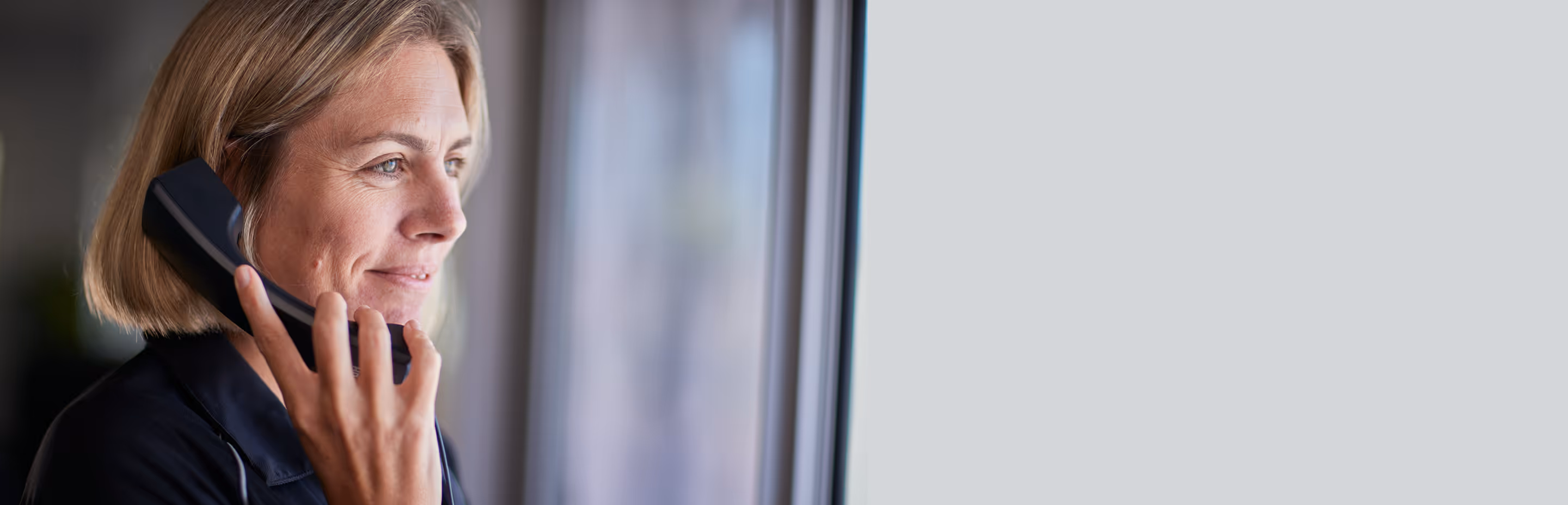 Woman with short blonde hair speaking on a corded telephone while looking out a window.
