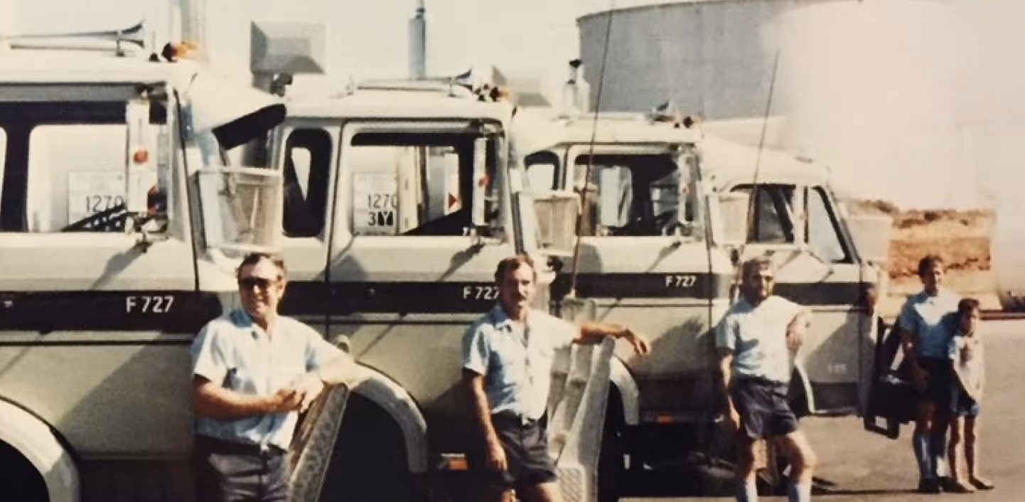 Four men in light shirts and dark shorts posing near and in front of three large white trucks with black stripe and markings F727.