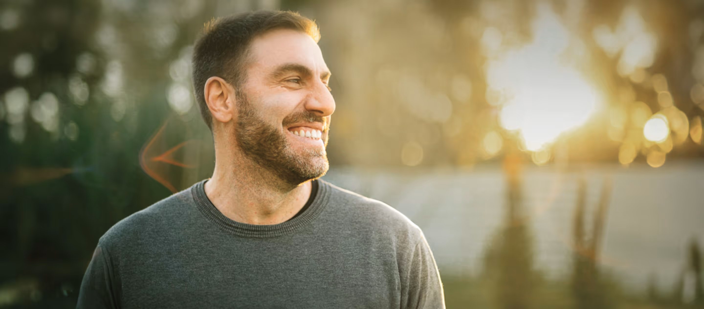 Smiling bearded man in a gray sweater looking to the side with a sunlit blurred background.
