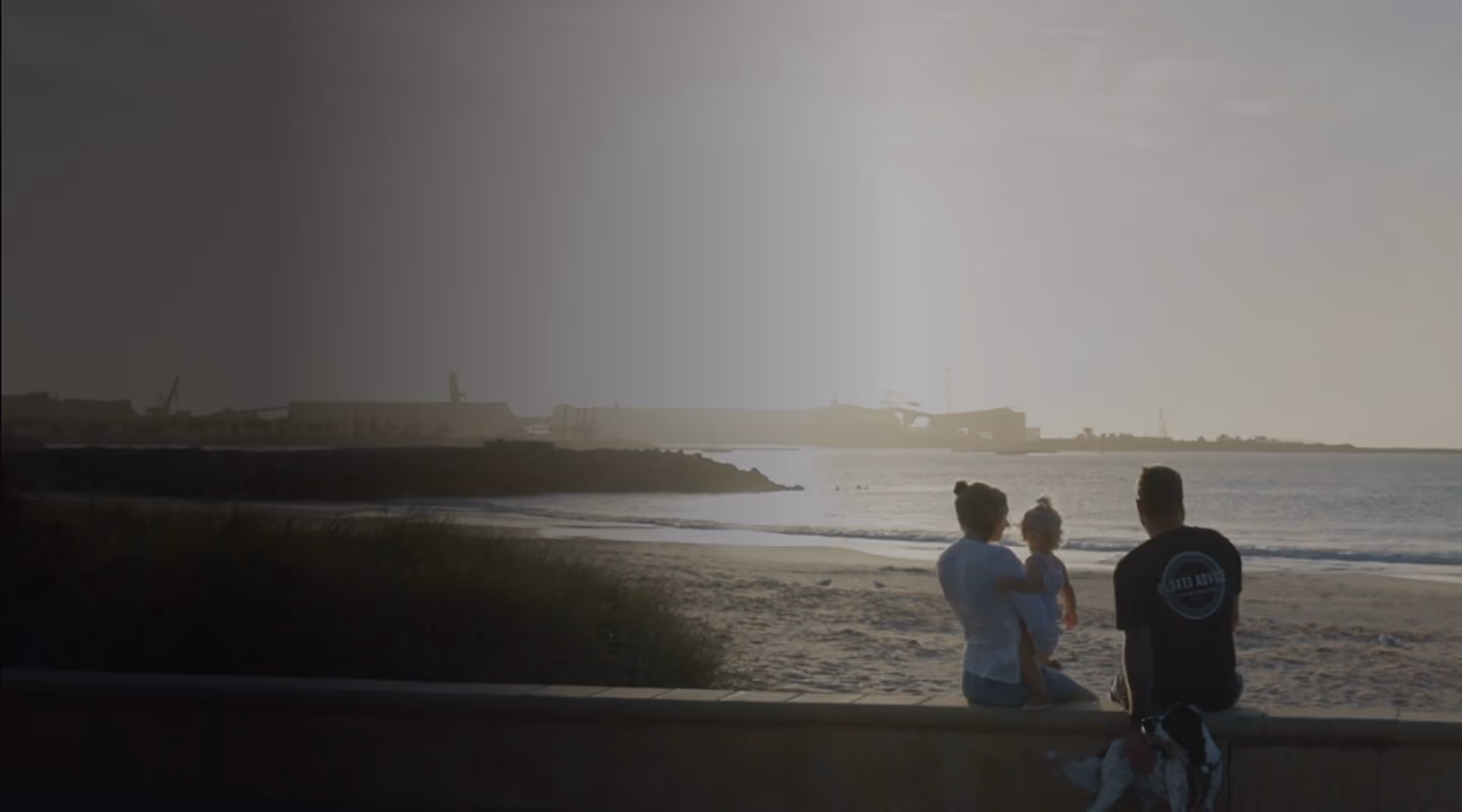 A family of three with a dog sitting on a low wall, watching the beach and ocean at sunset.