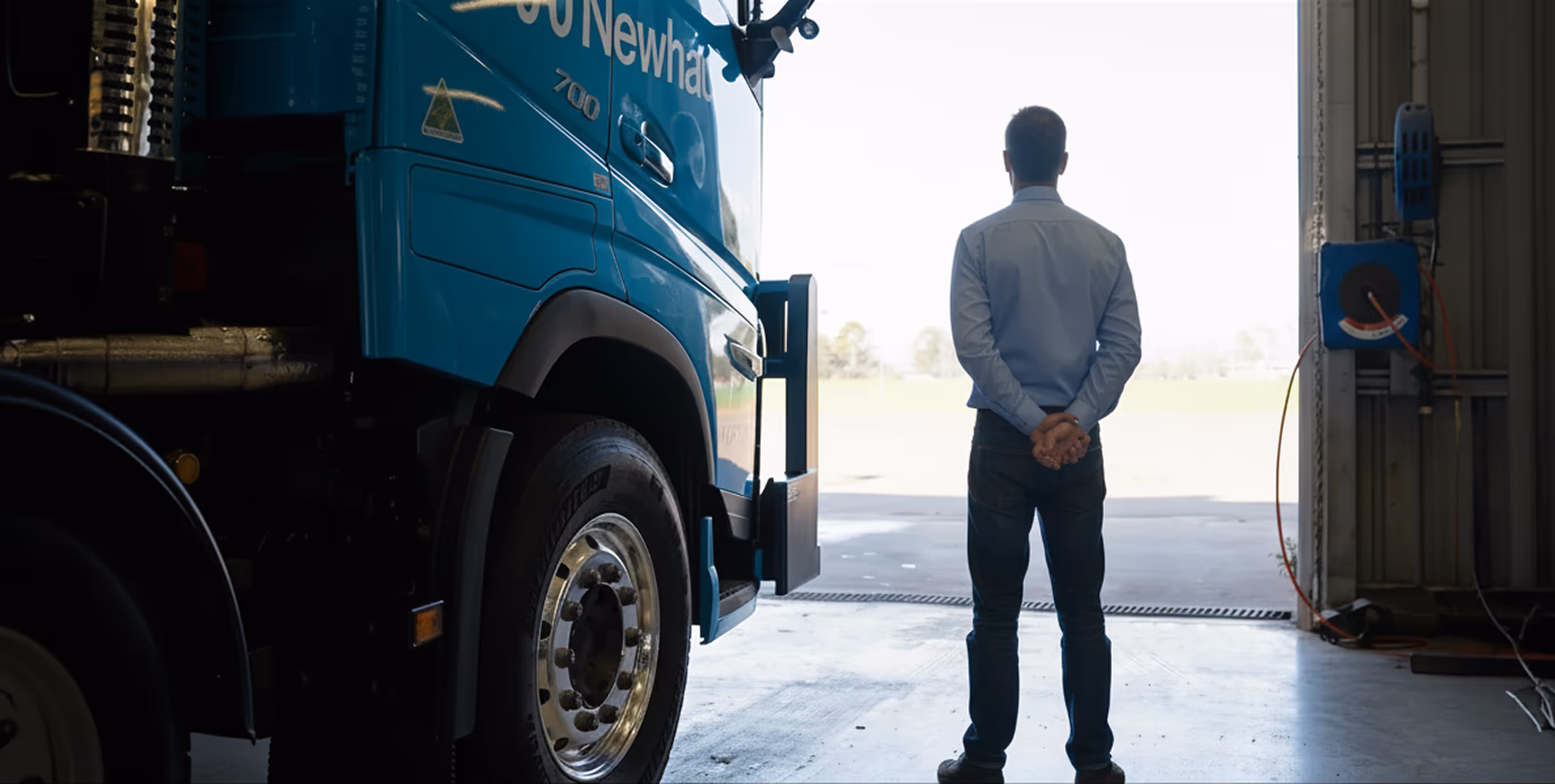 Man in a blue shirt standing with hands behind back, facing away near a large blue truck inside a garage.