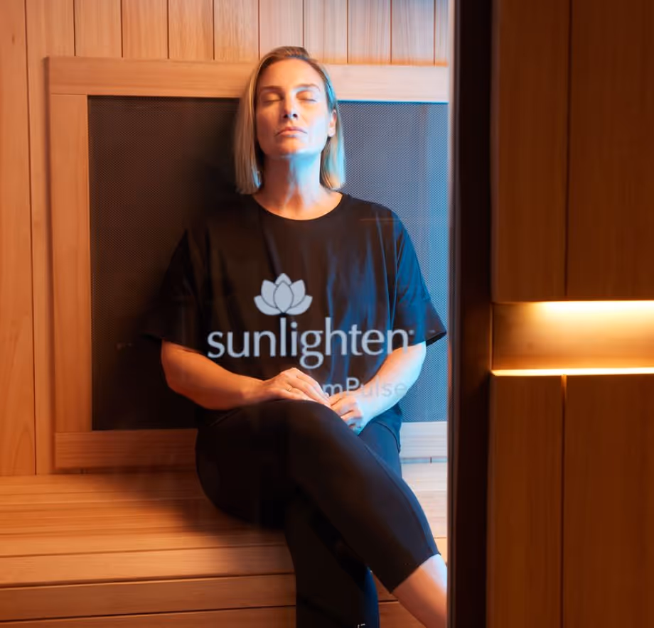 Woman with closed eyes sitting cross-legged on a wooden bench in a sauna with warm lighting.