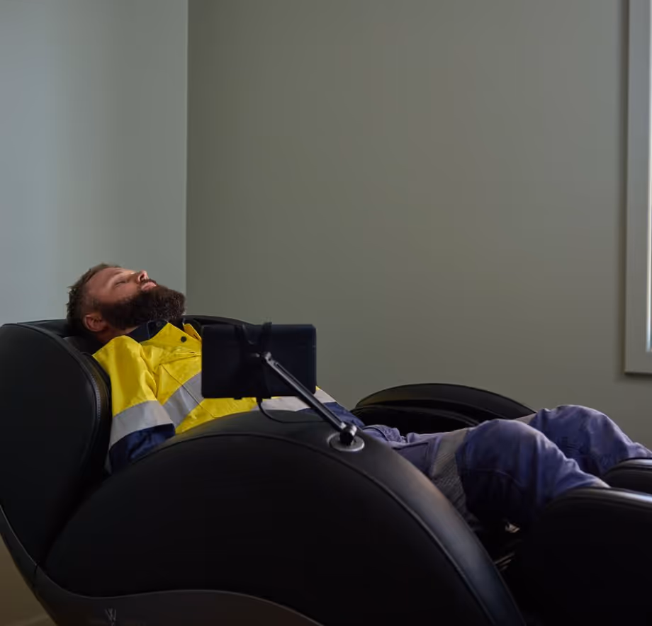 Man in a yellow and blue work uniform reclining in a black massage chair with eyes closed.