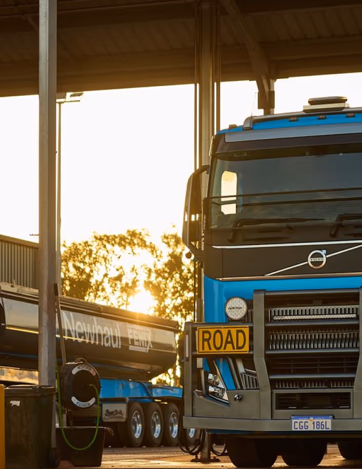 Front view of a blue Volvo road train truck parked at a station during sunset with a trailer labeled 'Newhaul Remix' in the background.