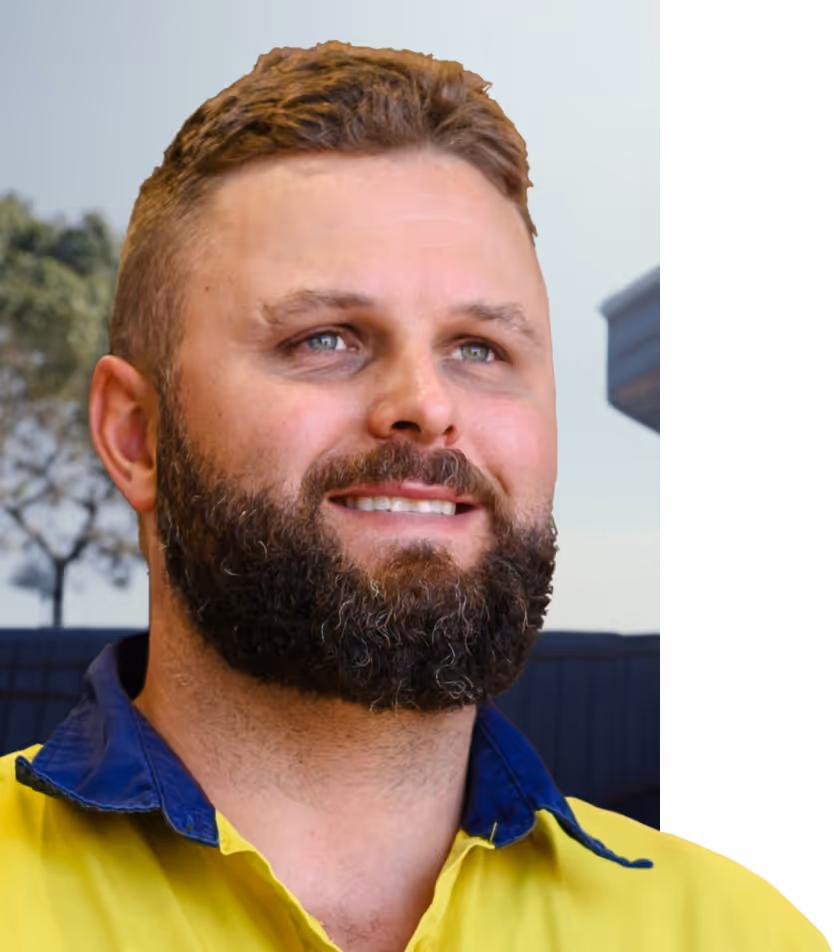 Bearded man with light blue eyes wearing a high-visibility yellow and blue work shirt, smiling and looking slightly to the side outdoors.