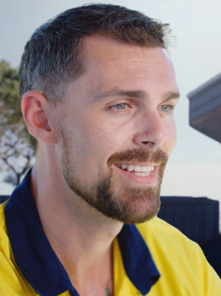 Close-up of a smiling man with short brown hair and a beard wearing a yellow shirt with a navy collar, outdoors.