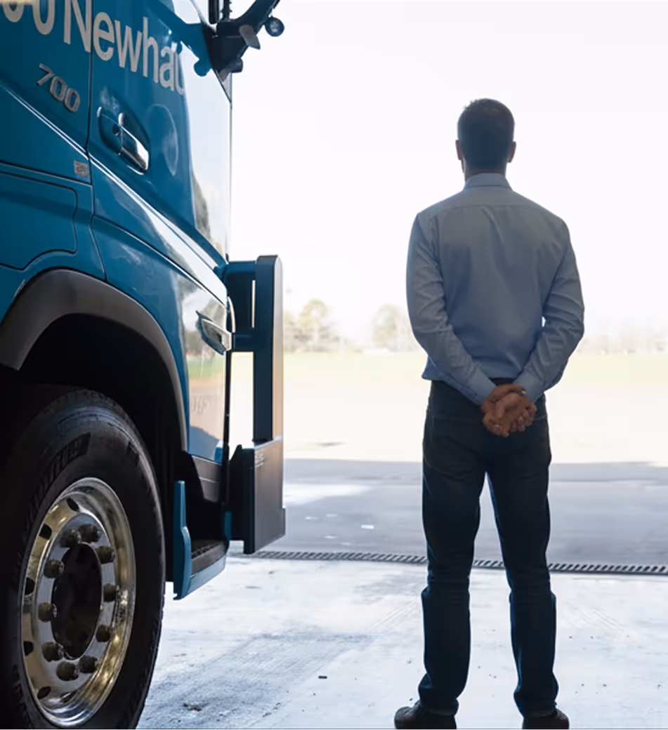 Man standing with hands clasped behind back, facing away, next to a blue Newhaul truck inside a Workshop.