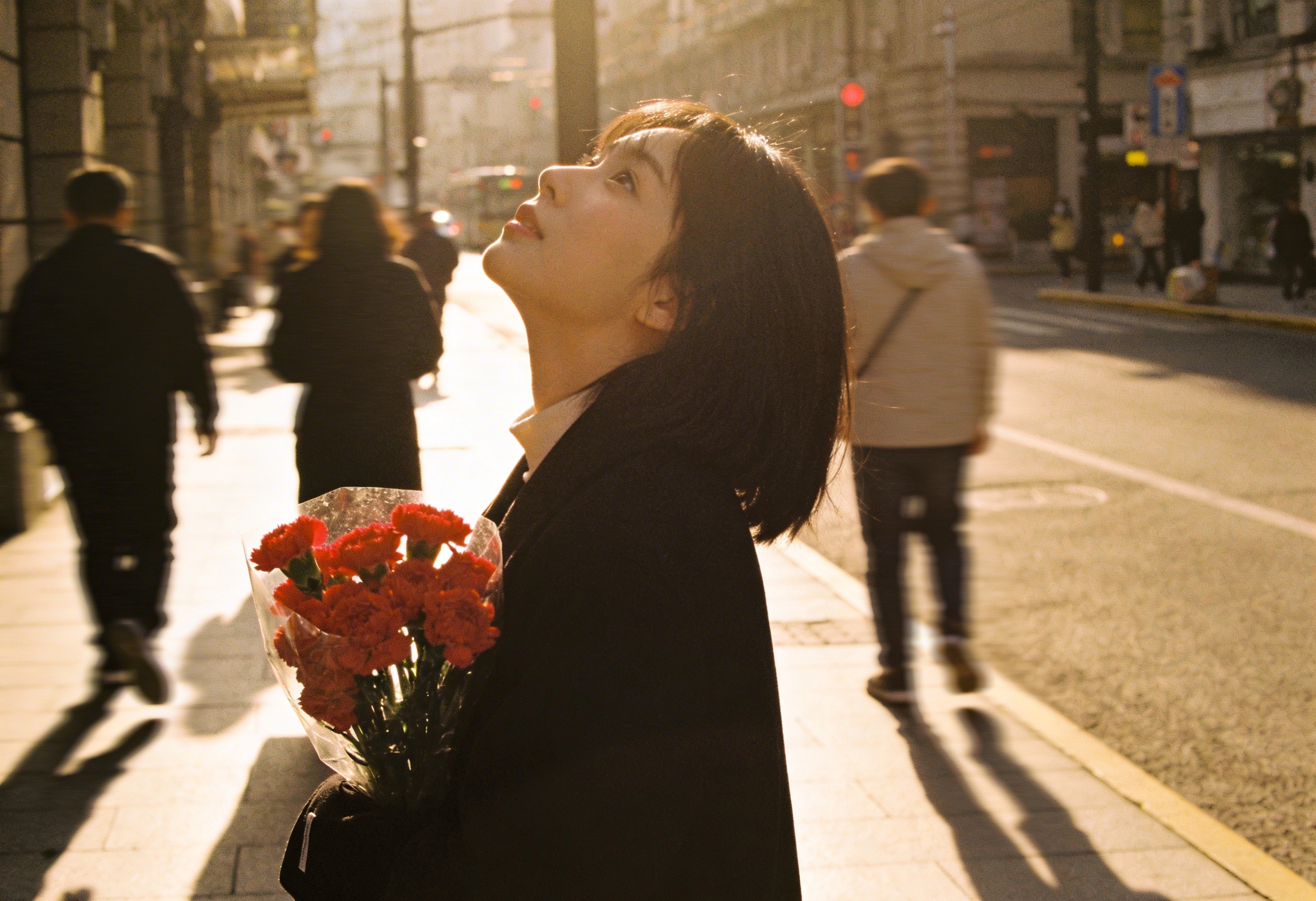 Young woman holding red carnations looks up on a sunlit city street with blurred pedestrians in the background.