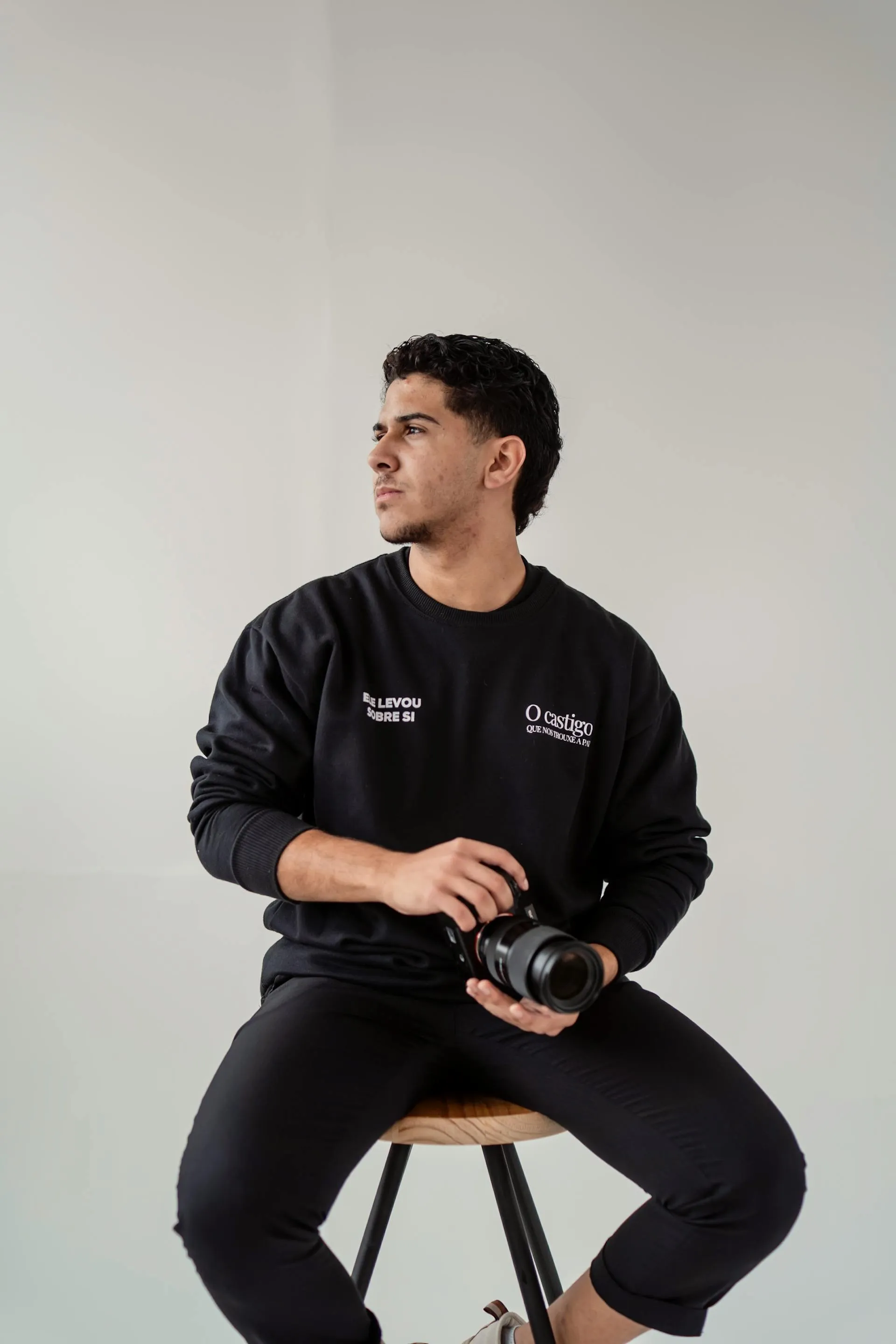 Young man in black sweatshirt holding a camera, sitting on a stool against a plain light background.