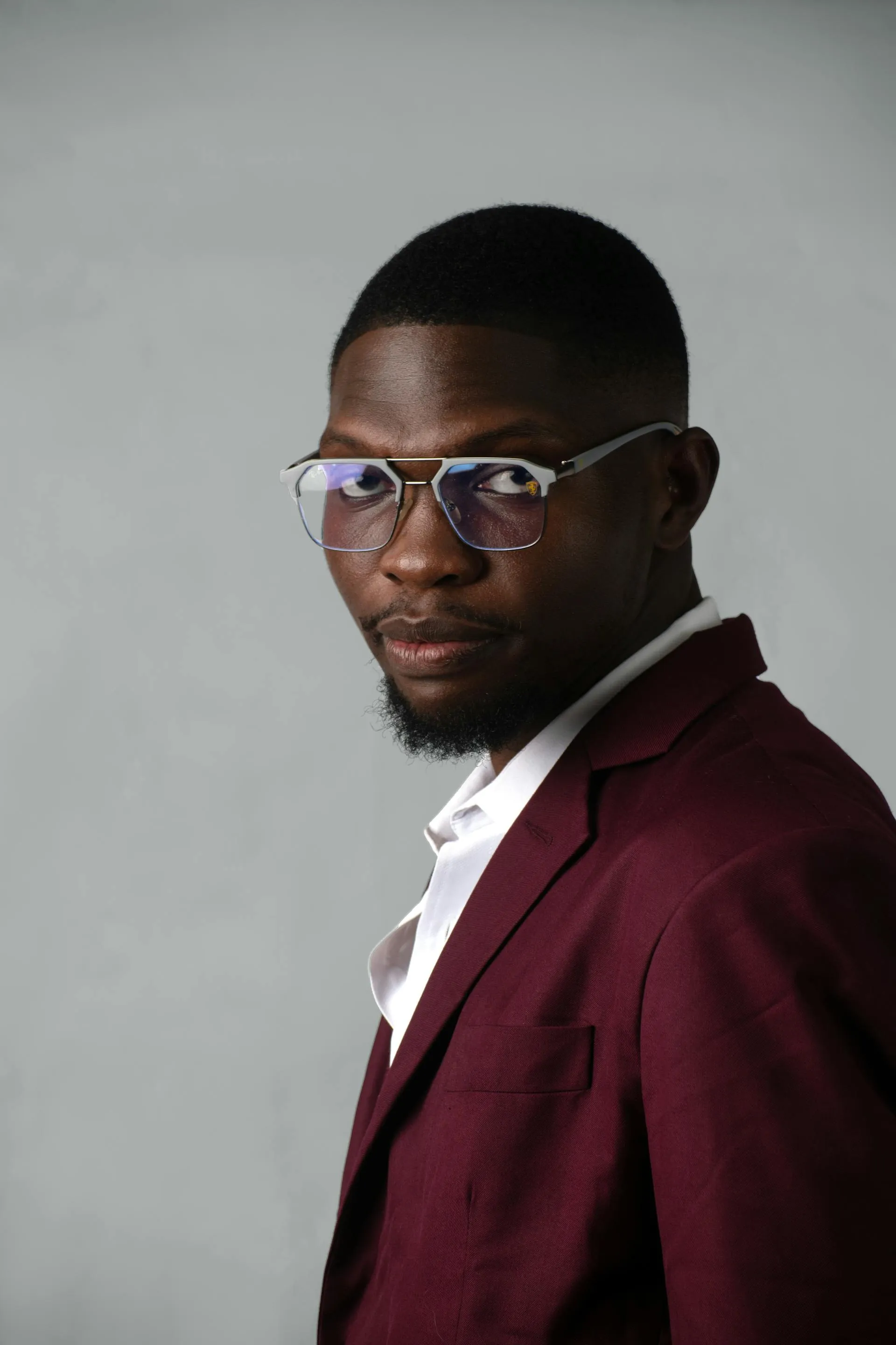 Young Black man wearing glasses and a burgundy blazer over a white shirt against a gray background.