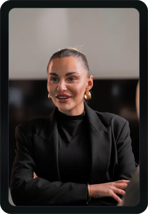 Eine Frau in schwarzem Blazer und schwarzen Ohrringen spricht und lächelt in einem professionellen Umfeld.