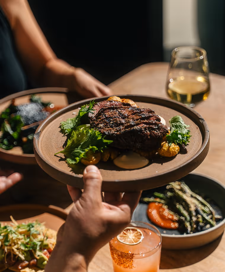 Server hands a plate of grilled ribeye steak with roasted potatoes and fresh herbs to a diner at a restaurant table.