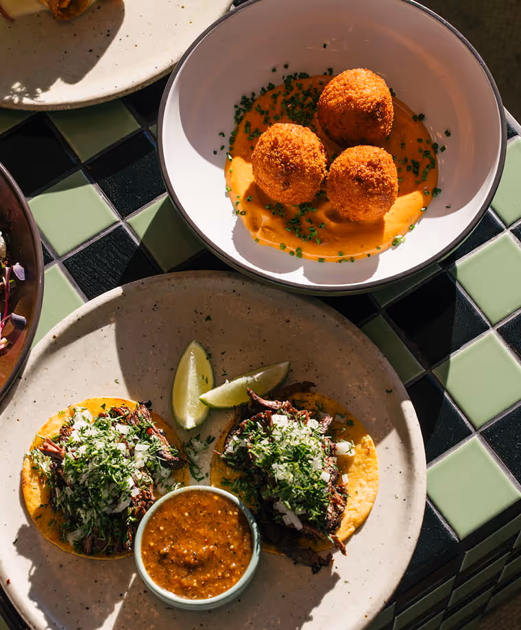 Crispy golden arancini balls in orange sauce and three barbacoa tacos topped with cilantro and onion on a checkered tile