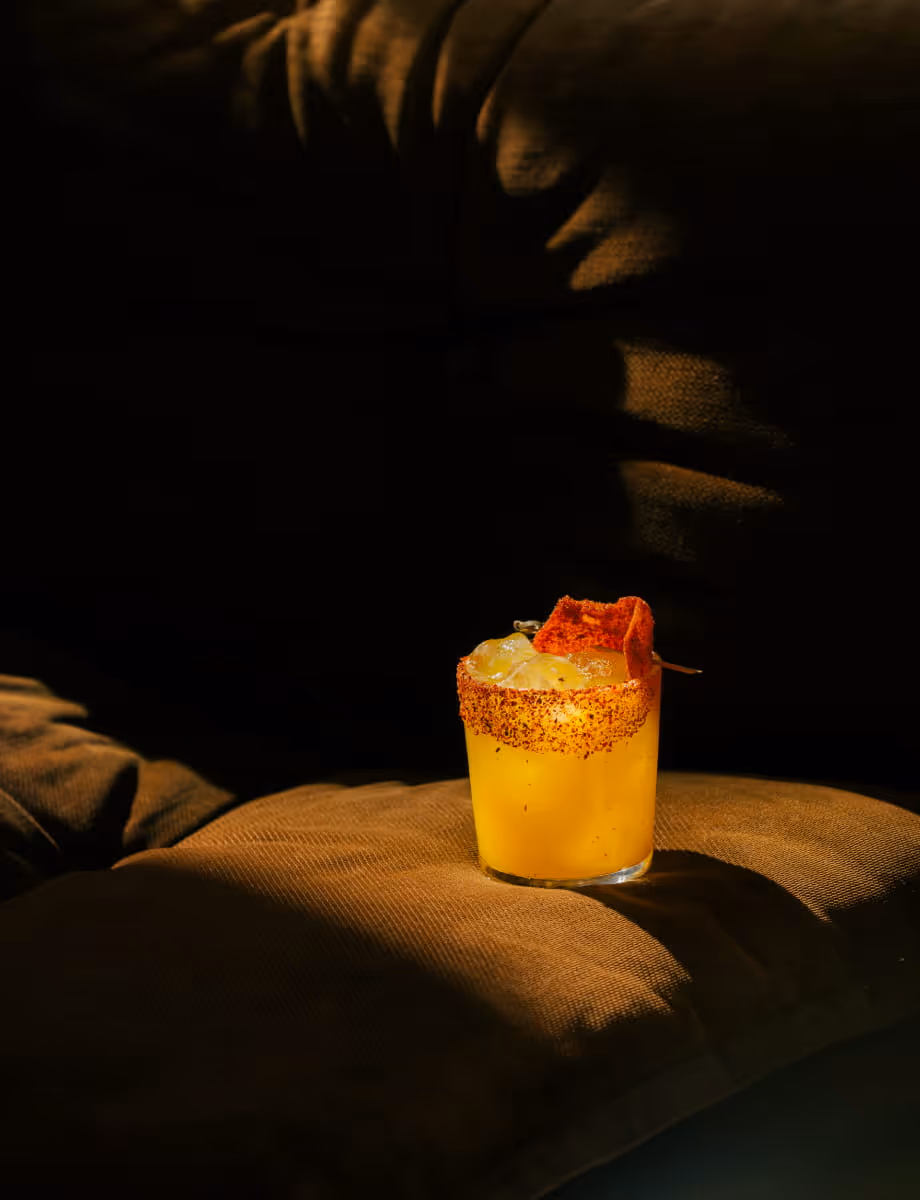 A vibrant mango margarita with a chili-salt rim and dried fruit garnish, served on a dark velvet cushion in moody lighting.