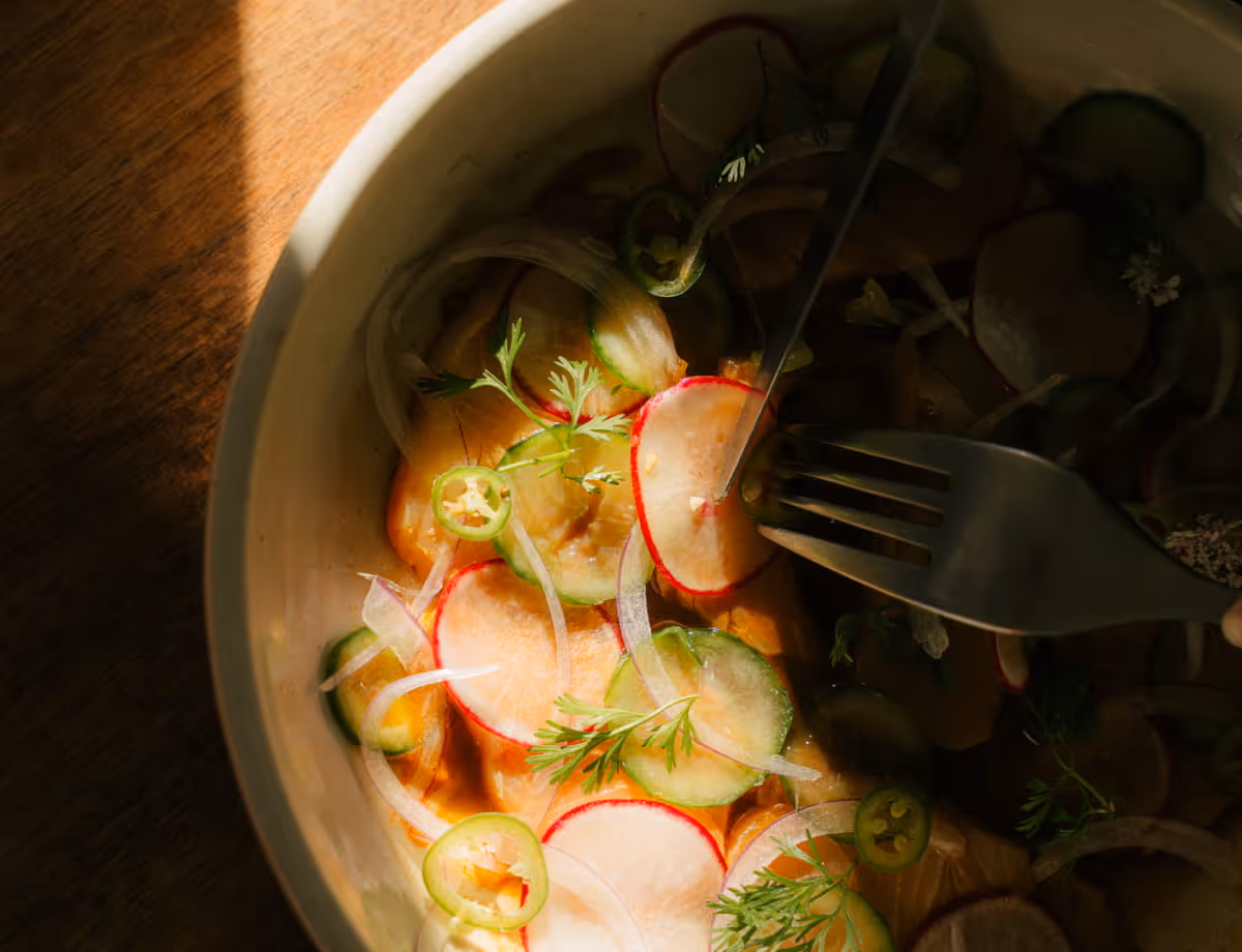 Fresh aguachile or ceviche with thinly sliced radishes, cucumbers, jalapeños, and herbs in citrus broth with a fork.