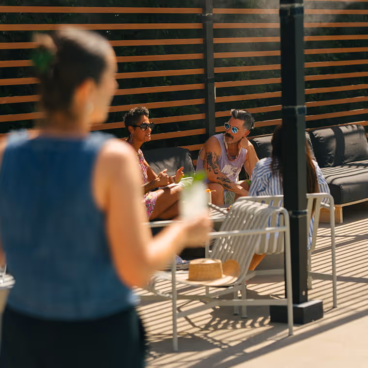 Friends relaxing on outdoor patio lounge furniture with drinks while a woman in denim approaches carrying a cocktail.