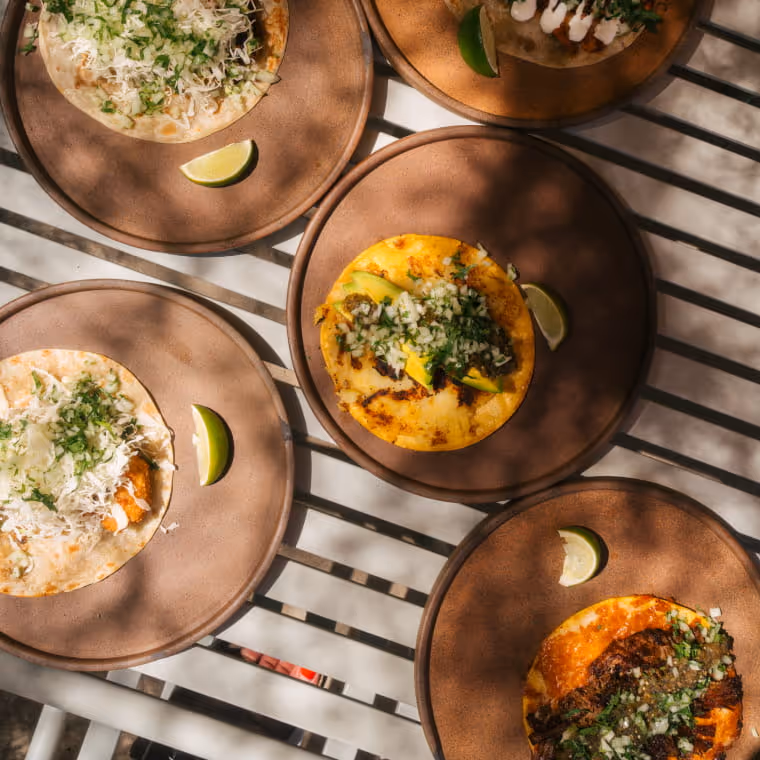 Overhead view of six tacos on clay plates garnished with cilantro, onions, avocado, and lime wedges on a metal table.