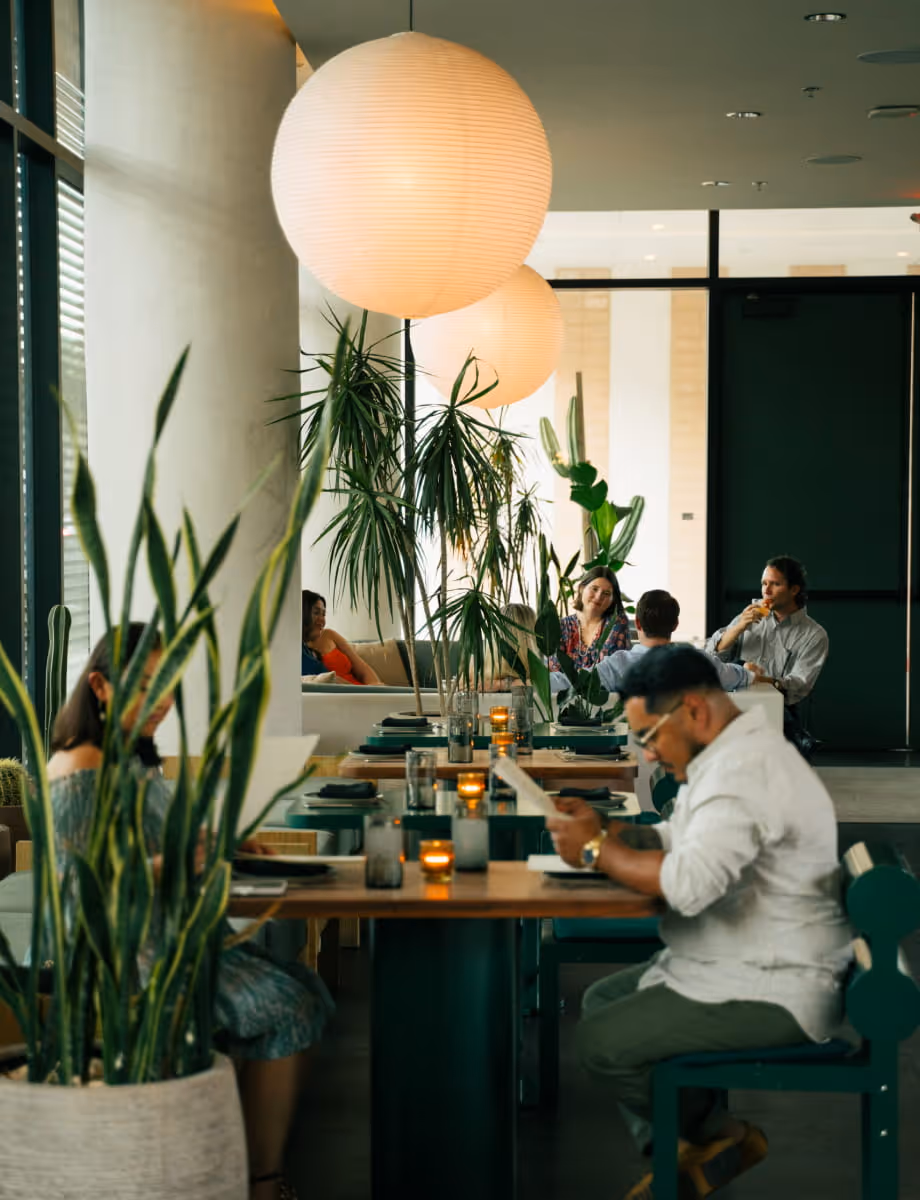 Stylish restaurant interior featuring tropical plants, paper globe pendant lights, and diners enjoying evening meals.