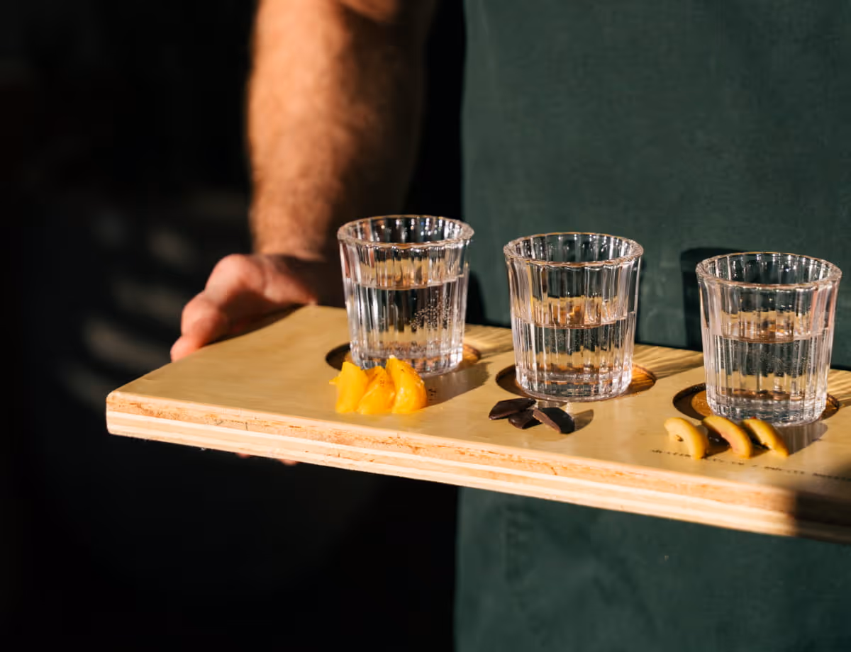 Server presenting a tequila or mezcal flight on a wooden board with orange slices and chocolate accompaniments.