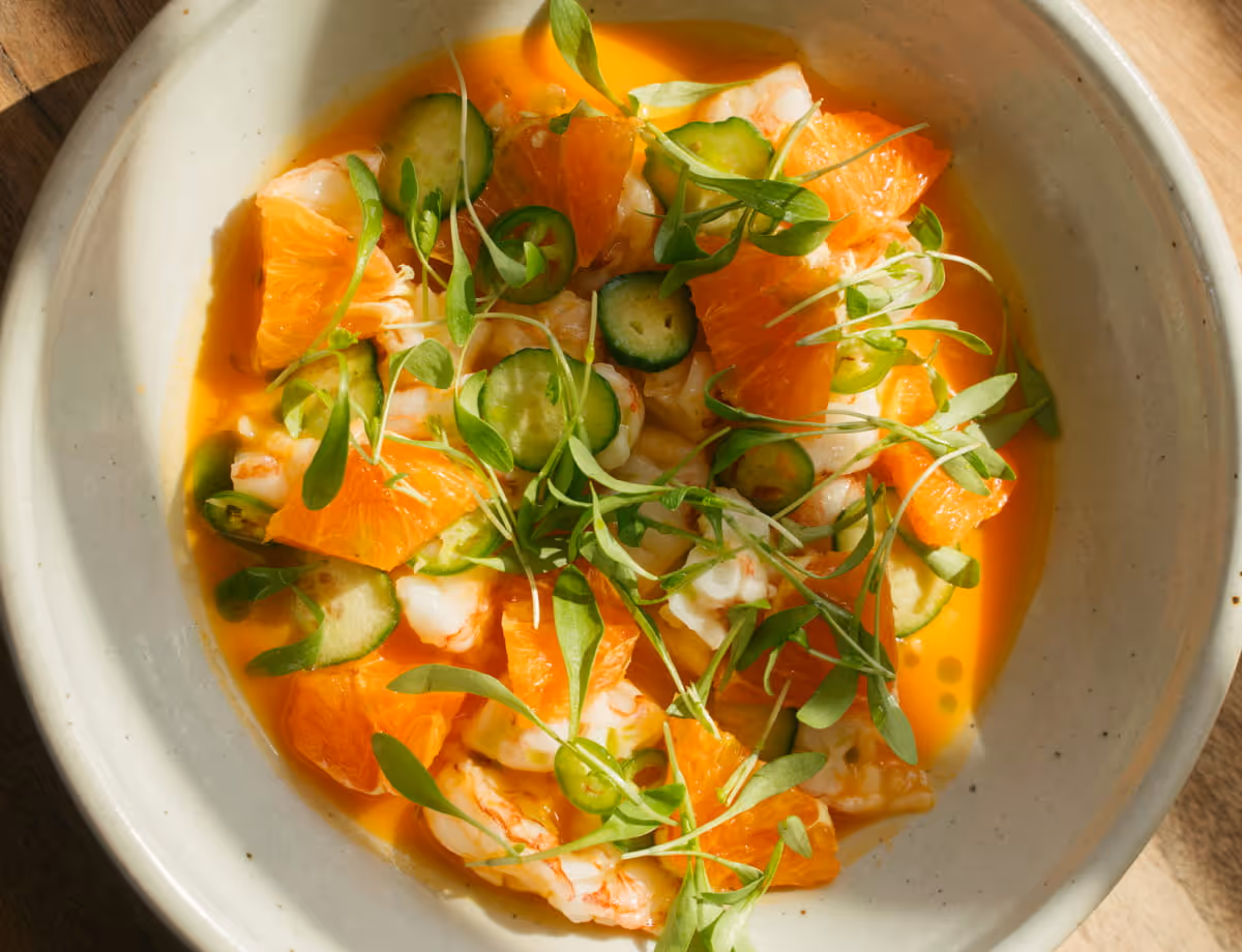 Fresh shrimp ceviche with citrus segments, cucumber slices, jalapeño, and microgreens in a vibrant orange broth.