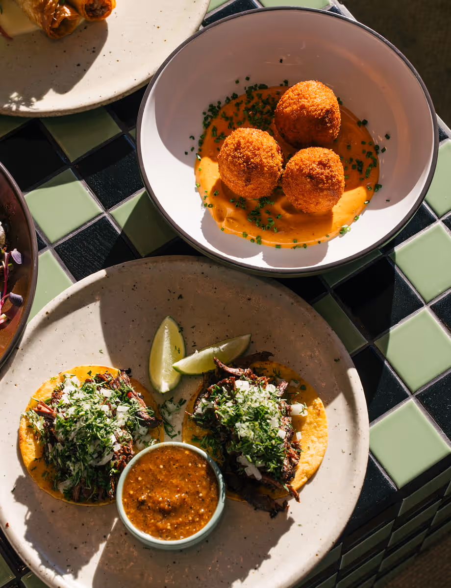 Crispy arancini balls in creamy orange sauce alongside beef tacos topped with cilantro and onion on a checkered tile table.