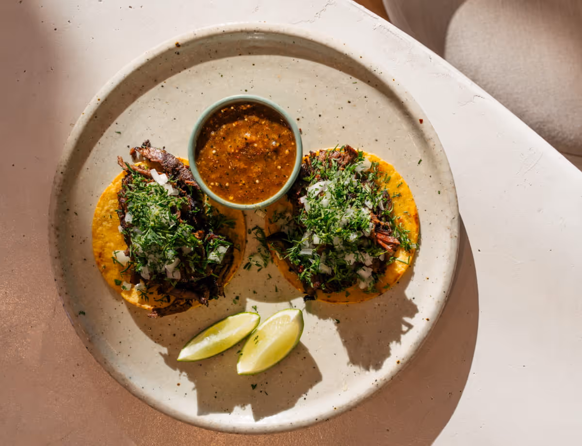 Two barbacoa tacos on corn tortillas topped with cilantro and onion, served with salsa and lime wedges on a speckled plate.