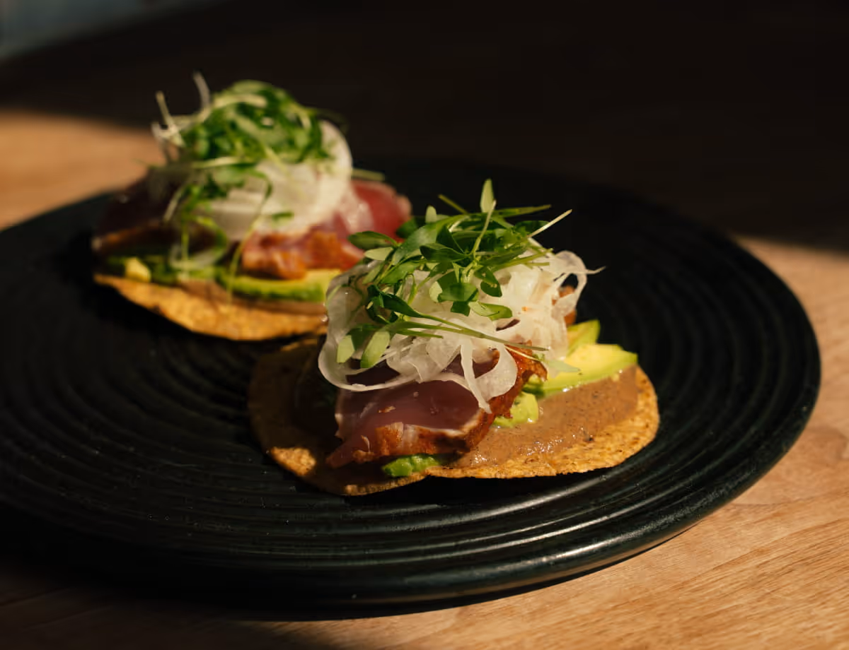 Two tuna tostadas topped with sliced avocado, shaved white onion, and microgreens served on a black ridged plate.