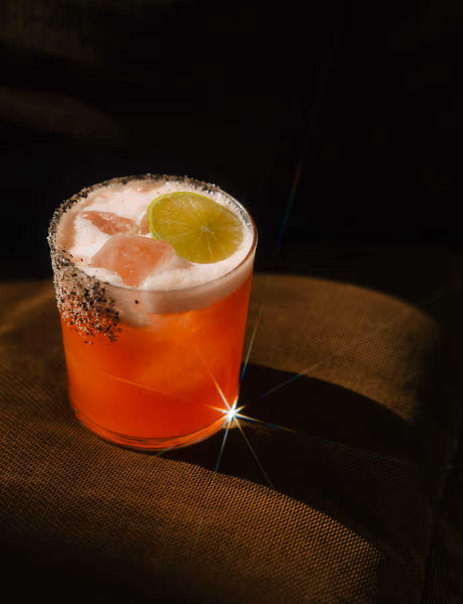 Vibrant orange margarita cocktail with a chili-salt rim, garnished with a lime wheel and ice cubes against a dark background.