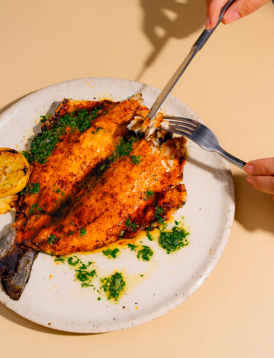 Whole grilled fish with paprika seasoning, herb gremolata, and charred lemon being cut with fork and knife on ceramic plate.