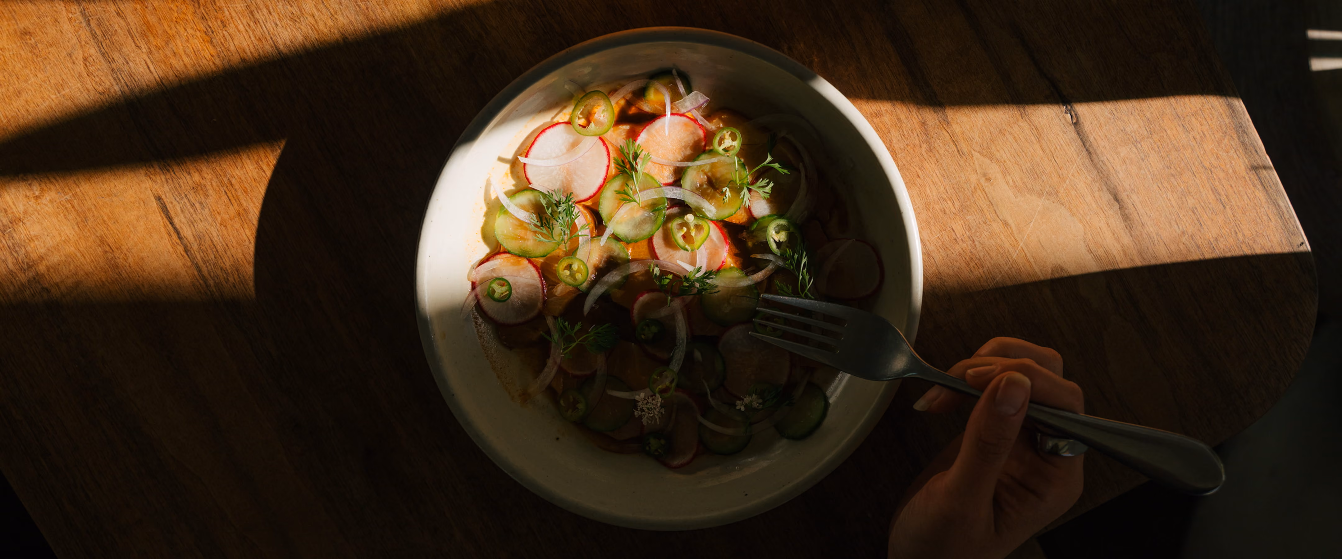 A hand holding a fork over a plate of fresh ceviche with radishes, cucumber, jalapeño, and herbs in golden sunlight