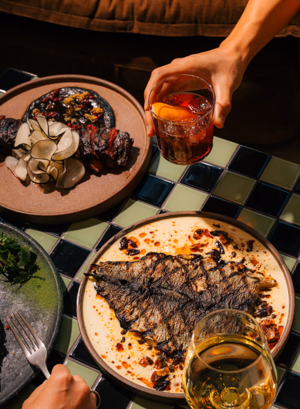 Overhead view of grilled whole fish and smoked meat dishes on a tiled table with a Negroni cocktail