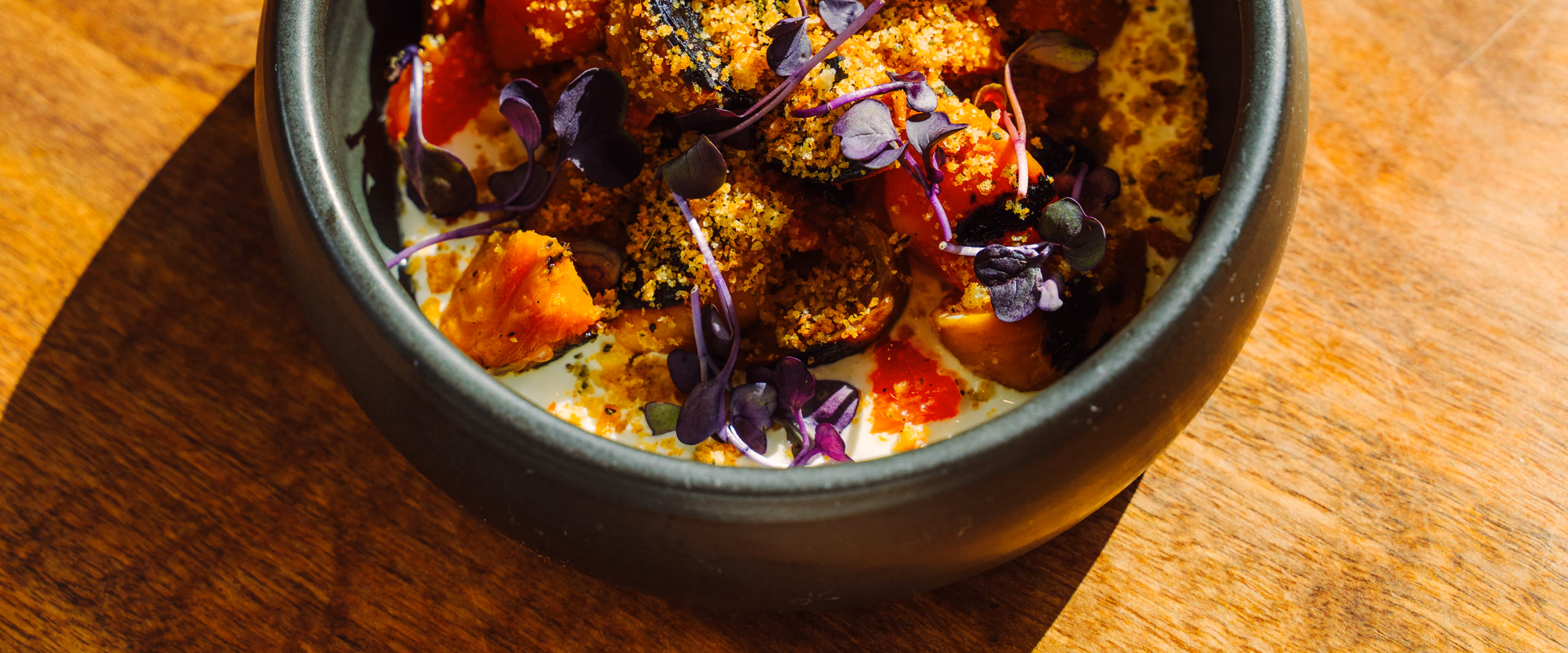A gourmet dish in a dark ceramic bowl featuring roasted vegetables, golden crumbs, and purple microgreens on a wooden table.