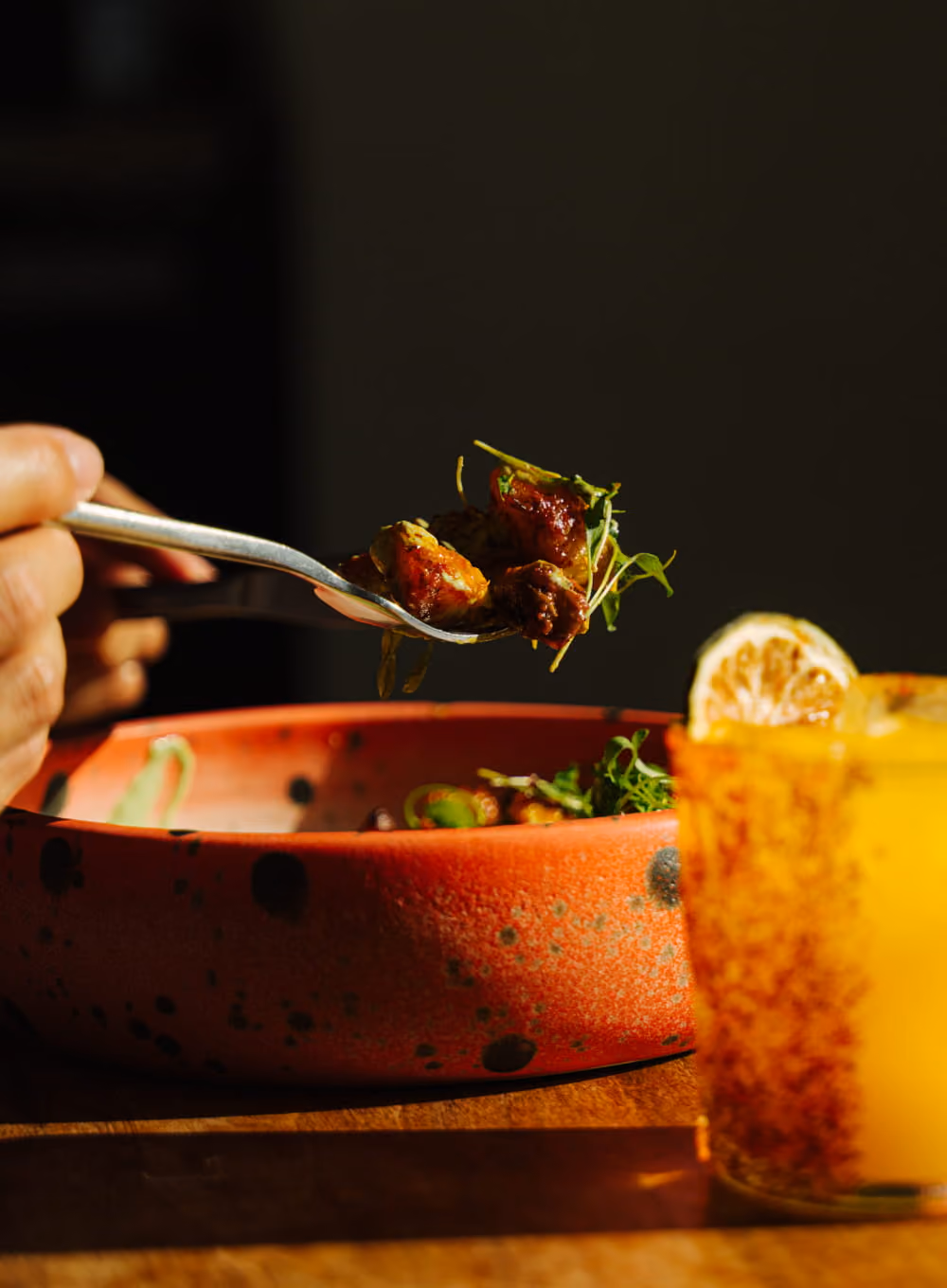A hand lifts a forkful of glazed meat and microgreens from a speckled red plate beside a citrus cocktail.