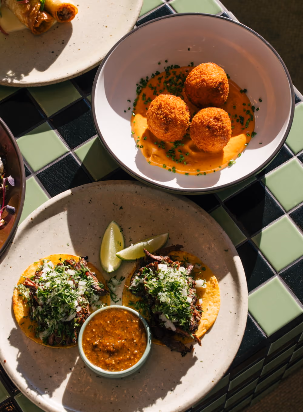 Overhead view of beef tacos with cilantro and lime on a plate beside crispy croquettes in orange sauce on a tiled table.