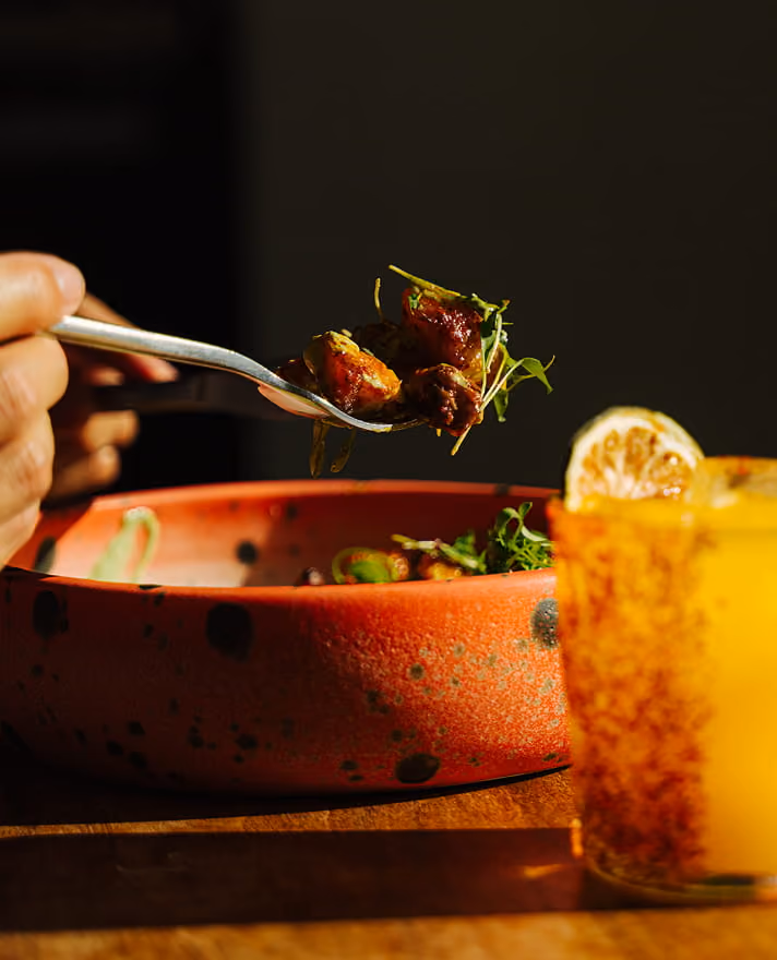 A hand lifts a forkful of glazed meat with microgreens from a speckled red bowl beside an orange cocktail with lemon.