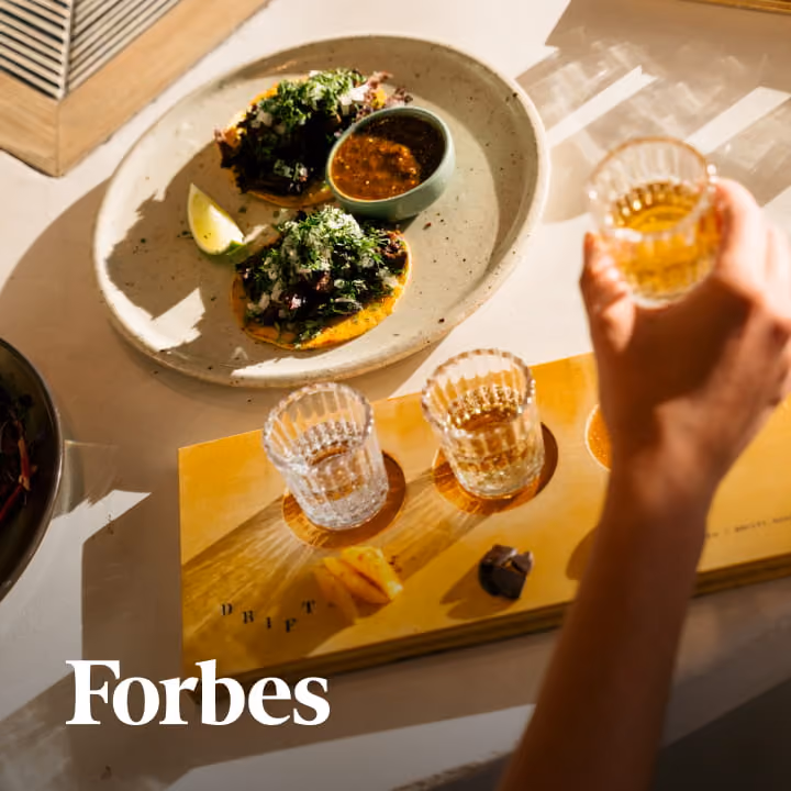 Tacos topped with cilantro and onion served with salsa alongside a tequila flight on a sunlit table, featured in Forbes.