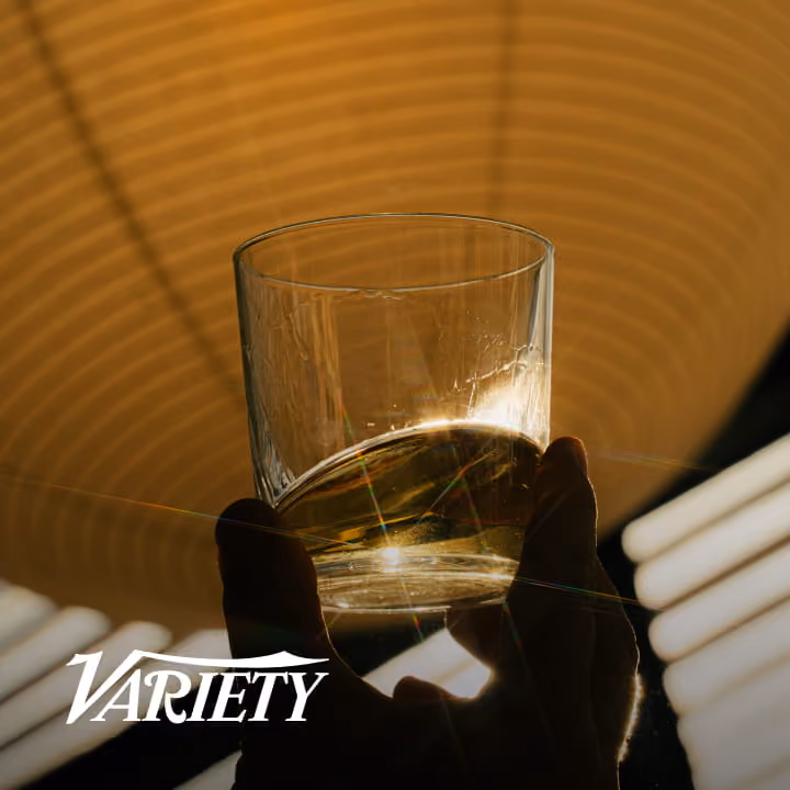 A hand holds a glass of golden whiskey up to warm backlight, creating a dramatic lens flare, with the Variety logo overlay.