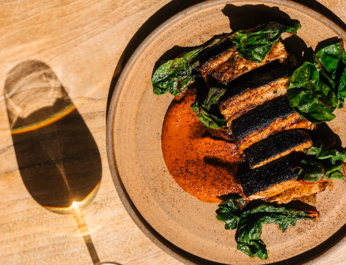 Sliced blackened pork belly served on a bed of sautéed chard with a smoky red sauce on a ceramic plate.